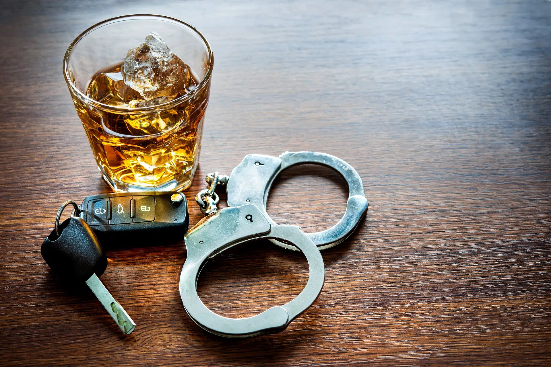 A glass of whiskey, car keys, and handcuffs on a wooden surface.